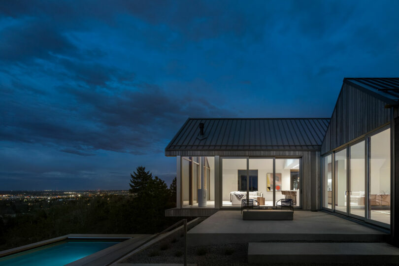 A modern villa with large windows and outdoor lighting at twilight, overlooking a cityscape, with a visible pool in the foreground.