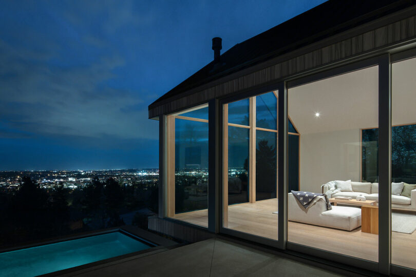 Villa H with large glass windows overlooking a city at night, featuring a well-lit interior living room and visible outdoor pool.