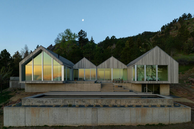 Modern villa with large glass windows and wooden siding, located in a forested area, under a twilight sky with a visible moon.