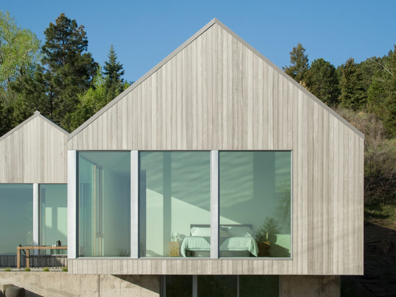 A modern villa with a gabled roof and large windows revealing a bedroom interior, set against a backdrop of green trees under a clear blue sky.