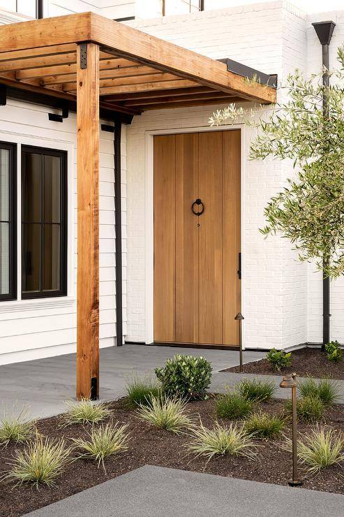 A white painted brick home features a pergola that sits over a plank front door with a bronze ring door knocker.