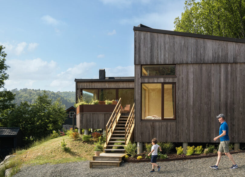 A modern wooden house with large windows and a raised deck sits on a grassy hill. A man and a child walk on a gravel pathway in front. Trees and a smaller house are in the background.