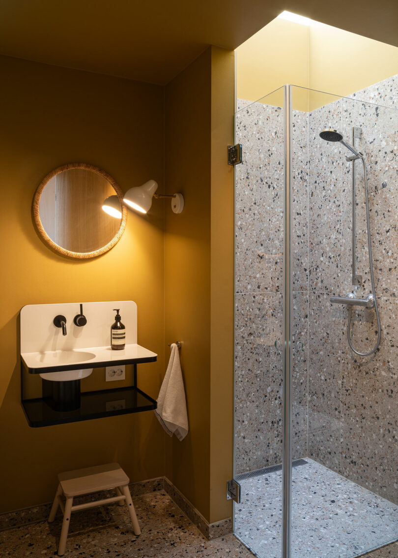 A modern bathroom with mustard yellow walls, a round mirror, a white sink with black fixtures, a wall-mounted light, and a glass-enclosed shower area with terrazzo tiles.