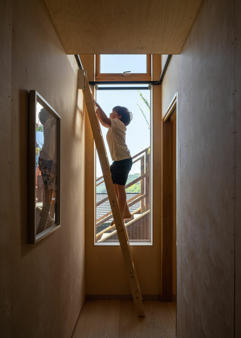 A child stands on a wooden ladder in a narrow hallway, reaching towards a high window. The hallway is illuminated by natural light from the window.