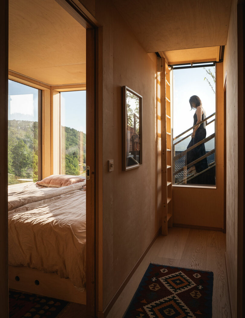 A small bedroom with a window view of greenery is shown through an open door. A person is ascending a staircase visible through a large window in the hallway. The interior is decorated with wall art and a rug.