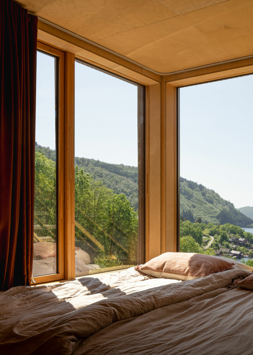 A sunlit bedroom with large corner windows showing a scenic view of green hills and a small town below.