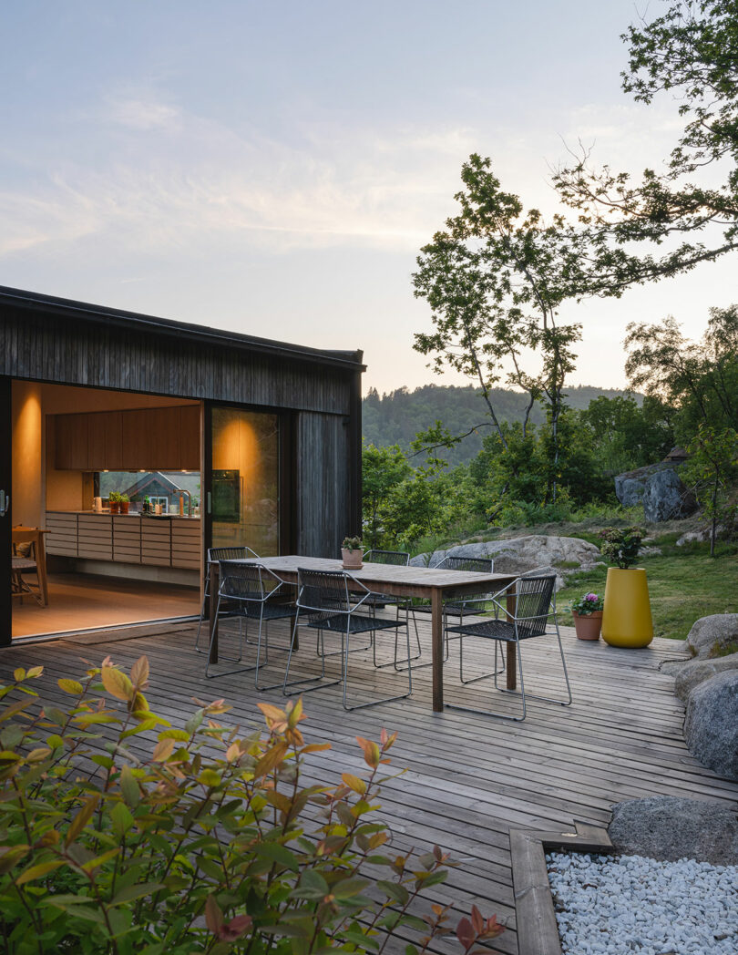 A modern patio with a wooden deck features an outdoor dining table and chairs. The patio is adjacent to a house with an open sliding glass door revealing a kitchen interior. Trees and greenery surround the area.