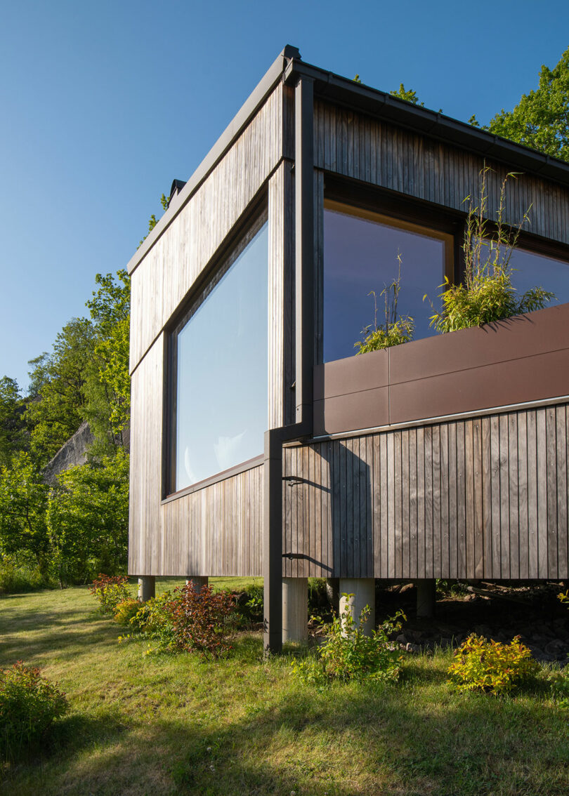 A modern wooden house with large windows and elevated on pillars, surrounded by green foliage and situated on a grassy slope.