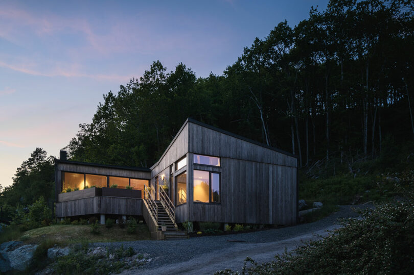 Modern house with large windows and wooden exterior, nestled on a hillside at dusk with surrounding trees and a gravel driveway.