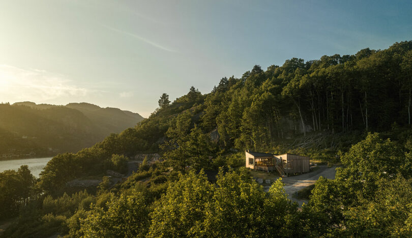 A wooden cabin is situated on a hillside surrounded by dense green forest, with a view of distant mountains and water under a clear sky.