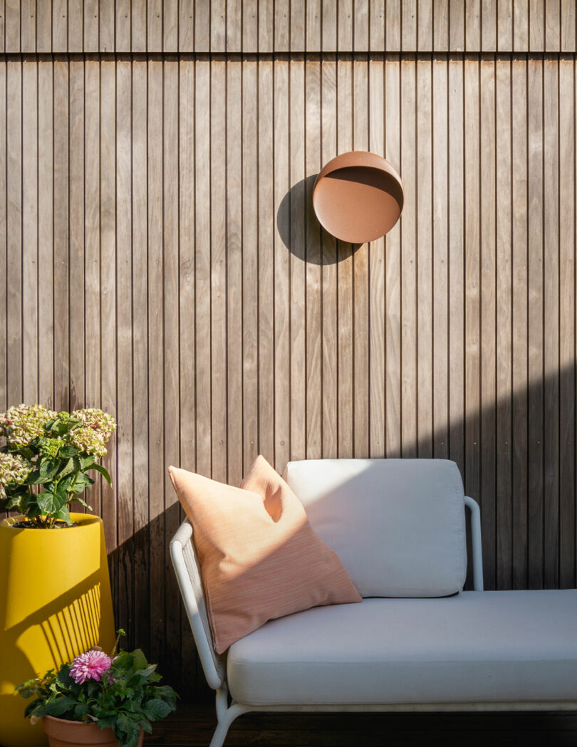 A white outdoor sofa with a peach pillow sits against a wooden slat wall. A yellow planter with flowers stands to the left, and a round, brown wall lamp casts a shadow on the wall above the sofa.