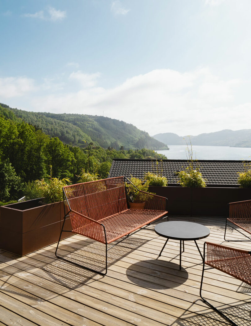 Wooden deck with wire furniture overlooks lush green hills and a water body in the distance under a partly cloudy sky.