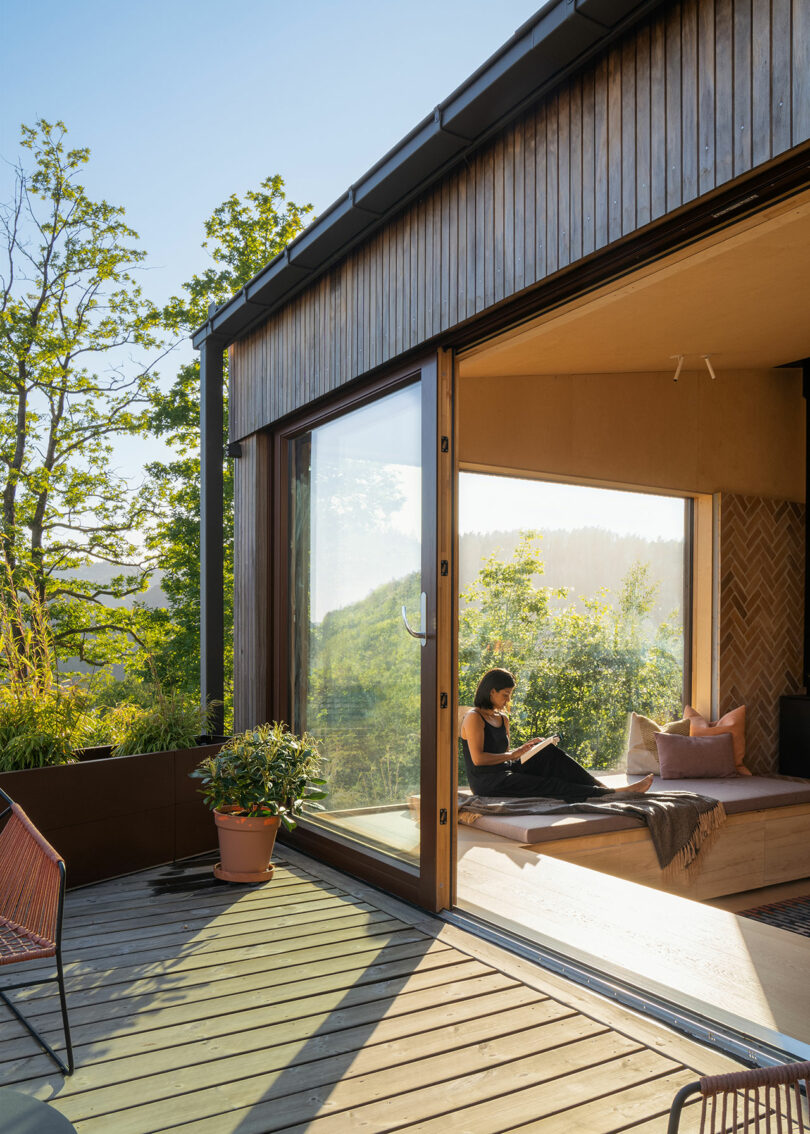 A person sits inside a modern house with a large glass sliding door, reading a book. The house overlooks a lush, green, outdoor landscape with trees and a deck area.