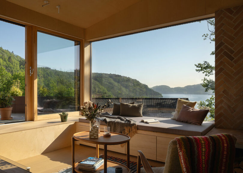 A cozy, sunlit room featuring large windows with a scenic view of mountains and a lake, containing a cushioned bench, coffee table, and assorted decor.