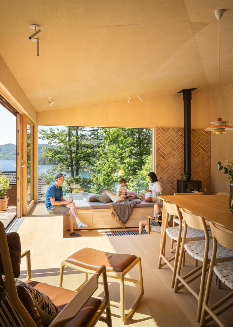 A family of three relaxes in a modern living room with wooden interiors, large windows, and a view of a lake and trees. The room features a small fireplace, chairs, a table, and decorative lighting.