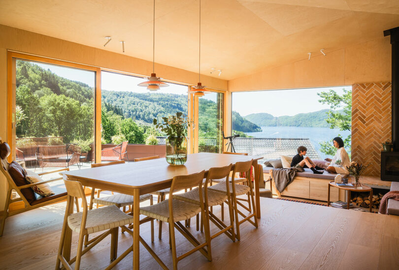 A spacious room with large windows offering a scenic view of a lake and mountains. The room has a wooden dining table, chairs, and two people sitting on a built-in sofa.