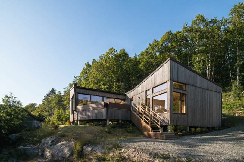 A modern wooden house with large windows, surrounded by trees, set on a slightly elevated plot with a gravel driveway leading to the main entrance.