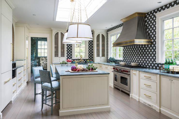 Hung beneath a skylight, white and gold pleated shade lights are positioned over a cream kitchen island accented with a gray marble countertop seating blue bamboo stools facing a sink with a brass faucet.