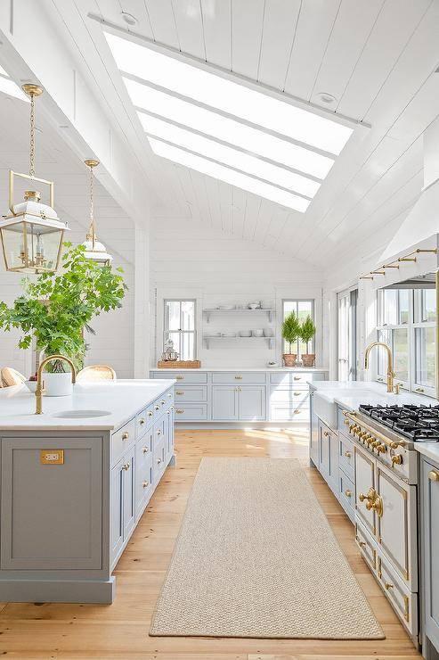 A skylight is fitted to a white plank vaulted ceiling is a gorgeous kitchen boasting white and gold carriage lanterns fixed over a light blue island finished with a honed marble countertop and prep sink with an antique brass gooseneck faucet.