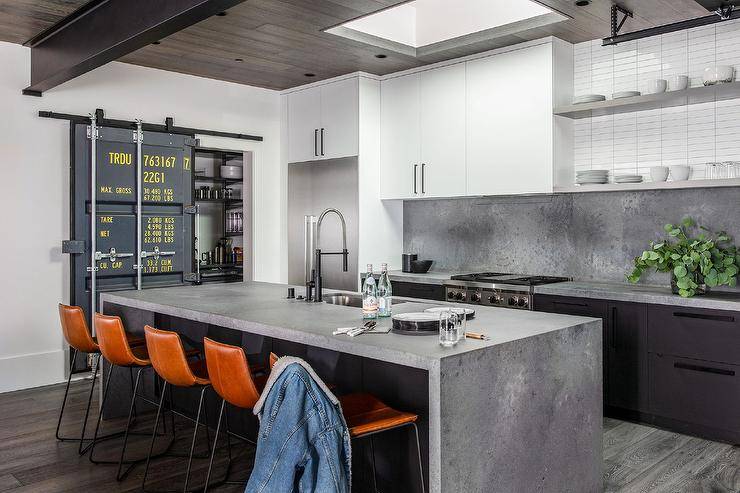 Stylish kitchen features a black center island complemented with a concrete waterfall edge countertop finished with a sink and a matte black pull out faucet. The faucet is mounted in front of brown molded leather counter stools illuminated by natural light from a skylight.