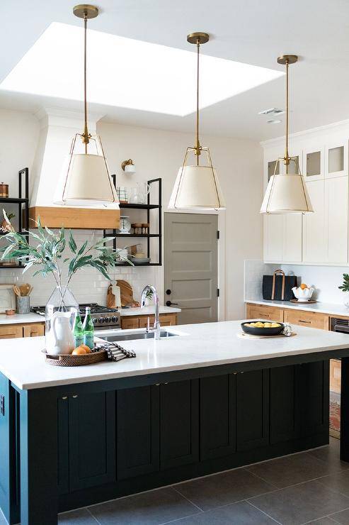 Bright kitchen features a large skylight over a black center island with brass knobs, topped in quartz, a sink with a nickel gooseneck faucet, lit by three brass hanging cage lights.