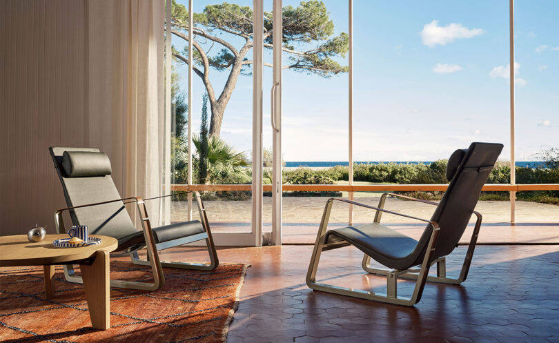 Two modern armchairs facing each other in a sunny room with large windows offering a view of the sea and trees. A table with a lamp and books is next to the chairs on a patterned rug.