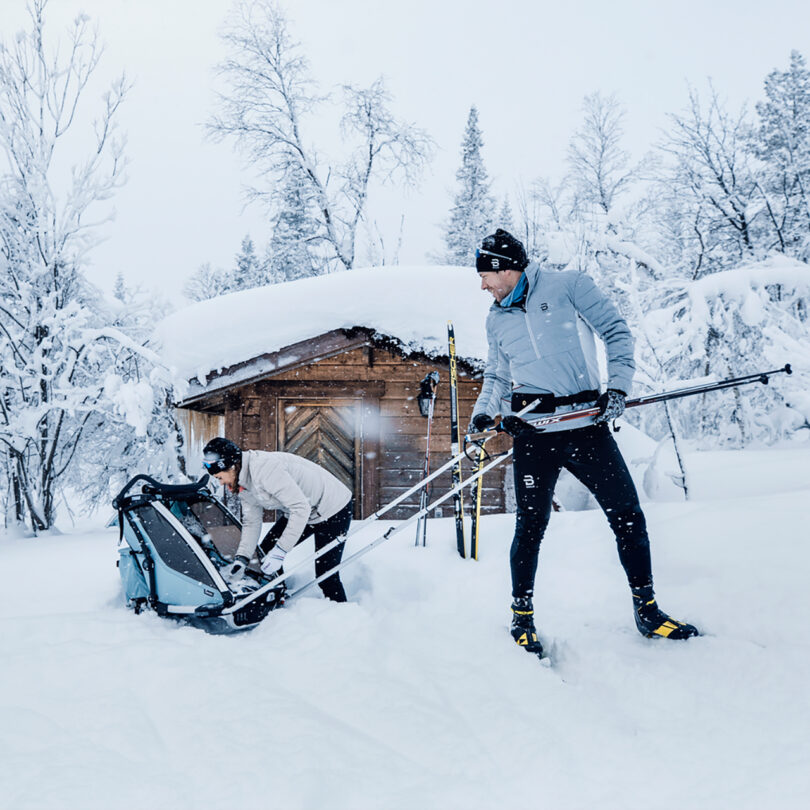 Two people outside in a snowy landscape with one person adjusting a ski stroller while the other holds skiing poles. A wooden shed is in the background, and both are dressed in winter sportswear.