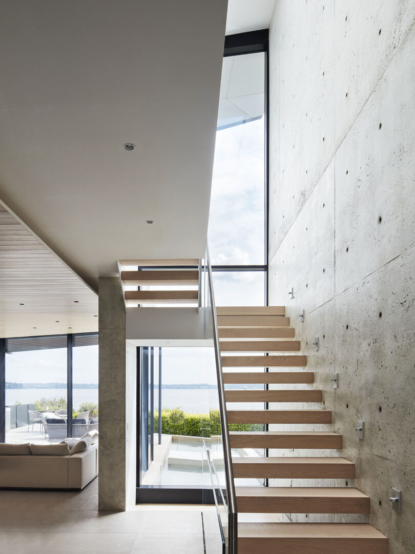 Minimalist interior with a wooden staircase, large windows, and concrete walls. The view outside shows a body of water and a partly cloudy sky. A seating area is visible to the left.