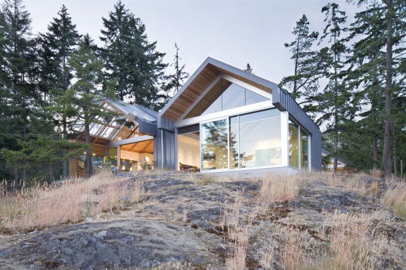 Modern house with large glass windows and sloped roof situated on a rocky, forested hillside. Trees and overcast sky are visible in the background.