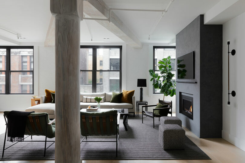 A modern living room with large windows, white walls, a fireplace, and green furniture, including a sofa, chairs, and a plant. The room features a pillar and a grey rug on the wooden floor.