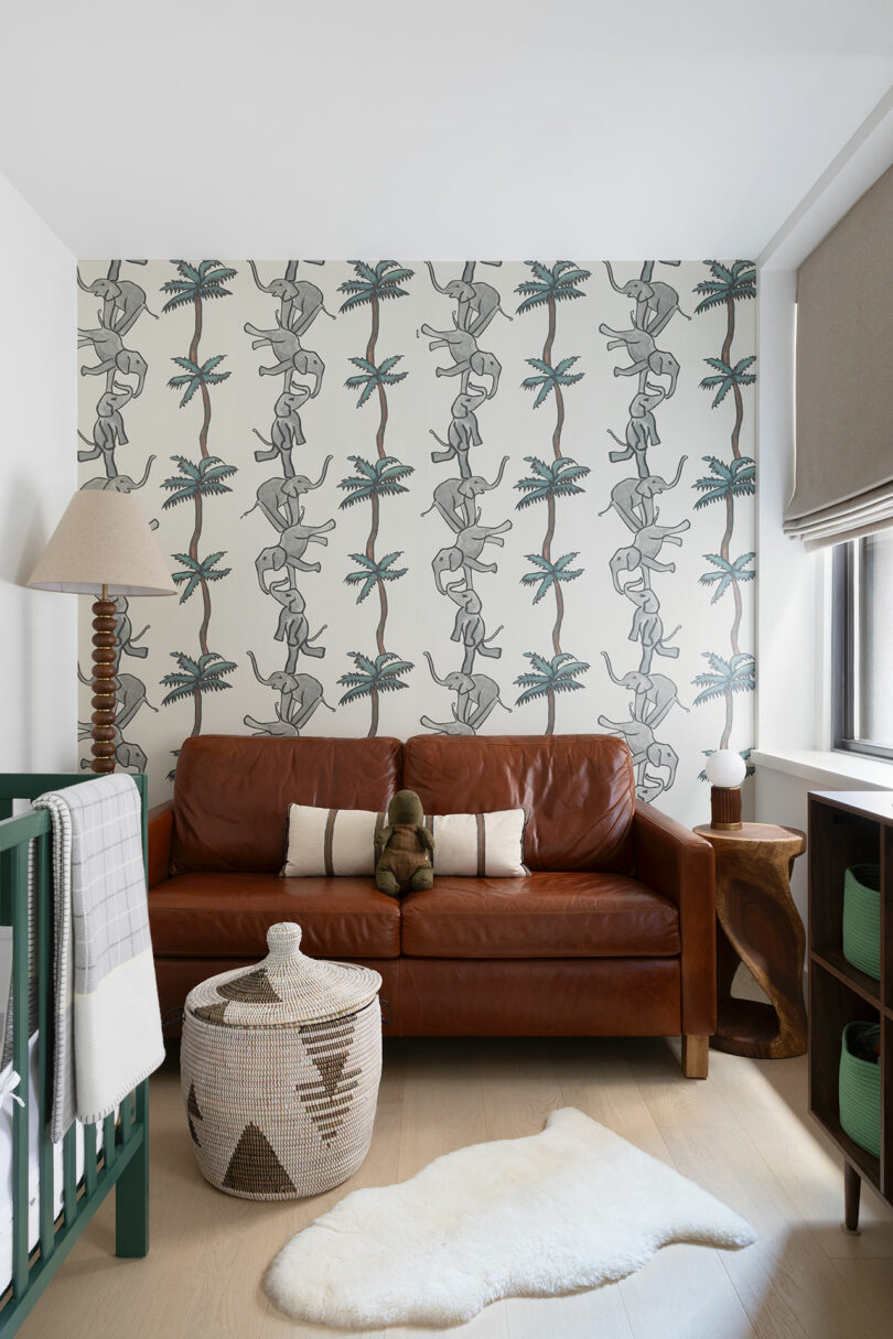 A cozy den features a brown leather sofa, a baby crib, a woven basket, and a palm tree-themed wallpaper. A plush toy sits on the sofa, and a sheepskin rug lies on the wooden floor.