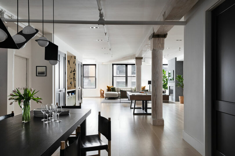 A modern open-concept living and dining area with a wooden table set with glasses, black chairs, white walls, exposed beams, large windows, and a seating area with a sofa and plants in the background.