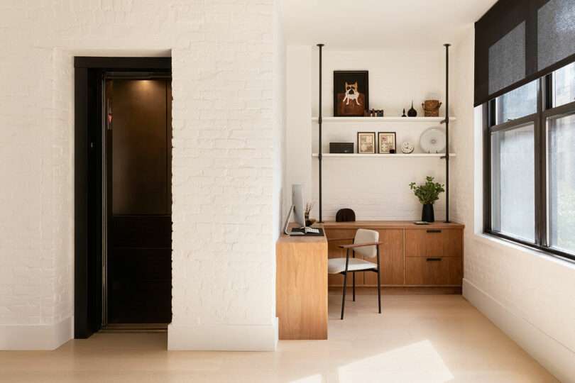 A minimalist home office with white brick walls, a wooden desk and shelving, a chair, and a window with a black blind. An elevator door is seen on the left.