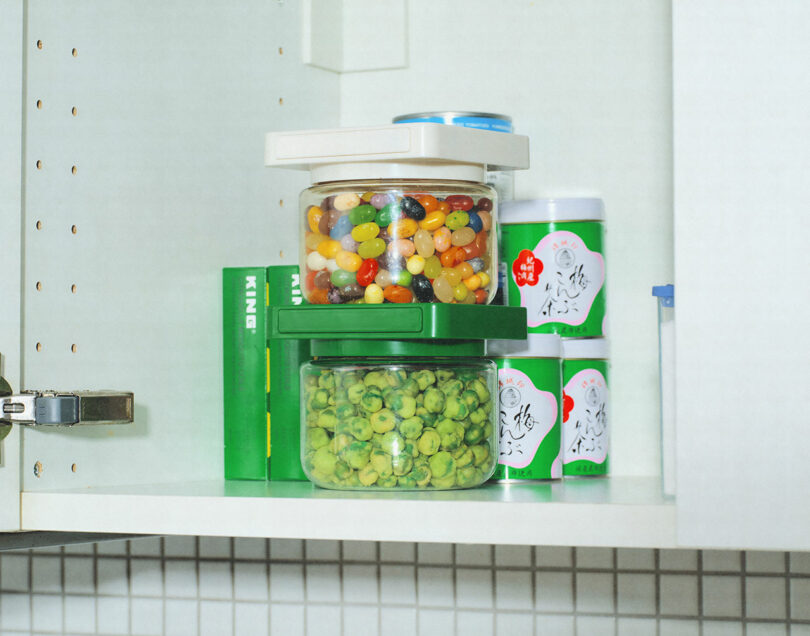 A kitchen cabinet shelf containing jars of jelly beans and dried peas, along with three cans of food products.