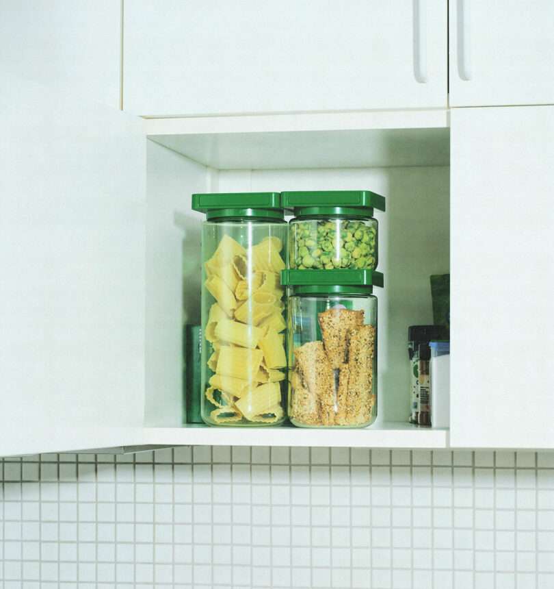 A kitchen cabinet with two shelves holding transparent containers filled with pasta, green peas, and granola bars, all with green lids.