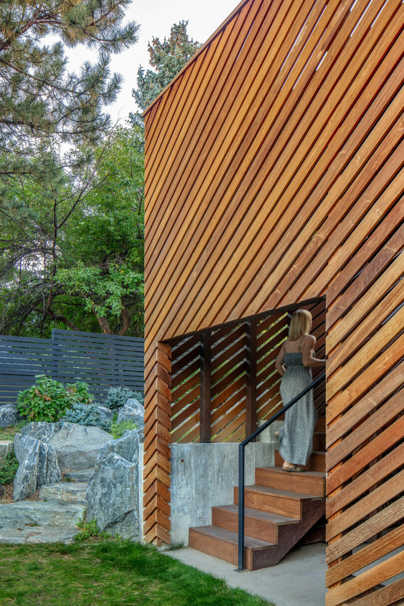 A person ascends an outdoor stairway built into a modern, diagonal wood-slatted structure, with a lush garden and rocks nearby.