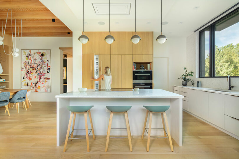 Modern kitchen with a central island featuring three bar stools, pendant lights, and wooden cabinets. A person is using the refrigerator. A dining area with colorful wall art and wooden ceiling is visible.