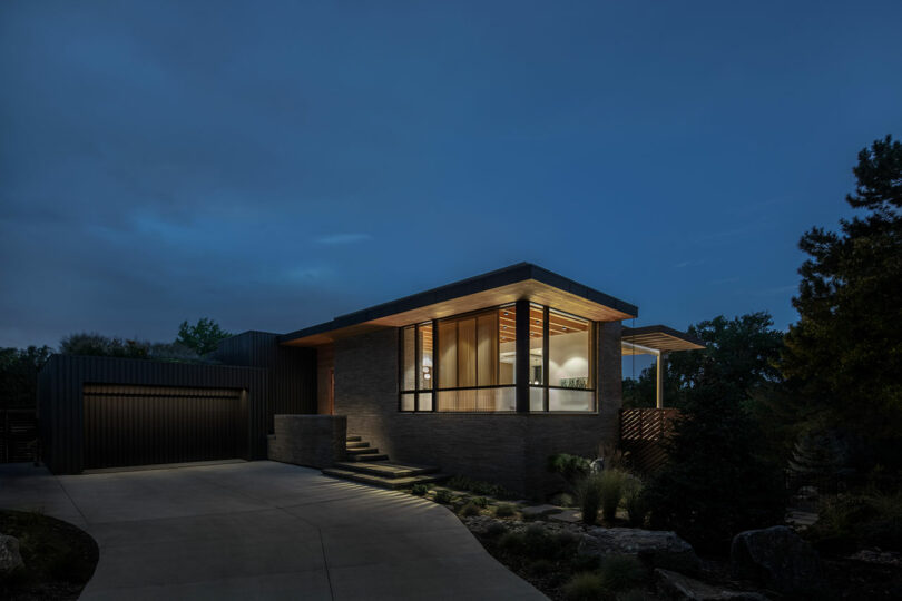 A modern house with large windows is illuminated at night. It has a flat roof and a pathway leading to the garage on the left side. Trees and bushes surround the property.