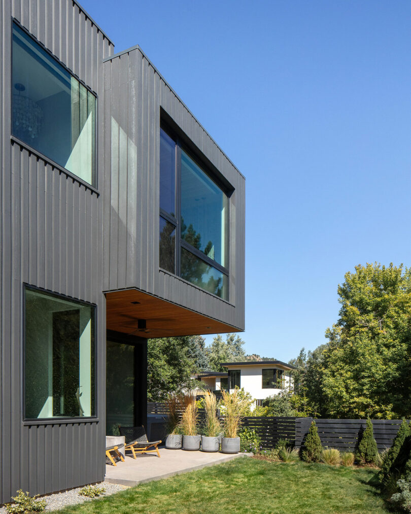 Modern two-story gray house with large windows, adjacent patio area with outdoor furniture and potted plants, and a fenced garden with green lawn and trees on a clear day.