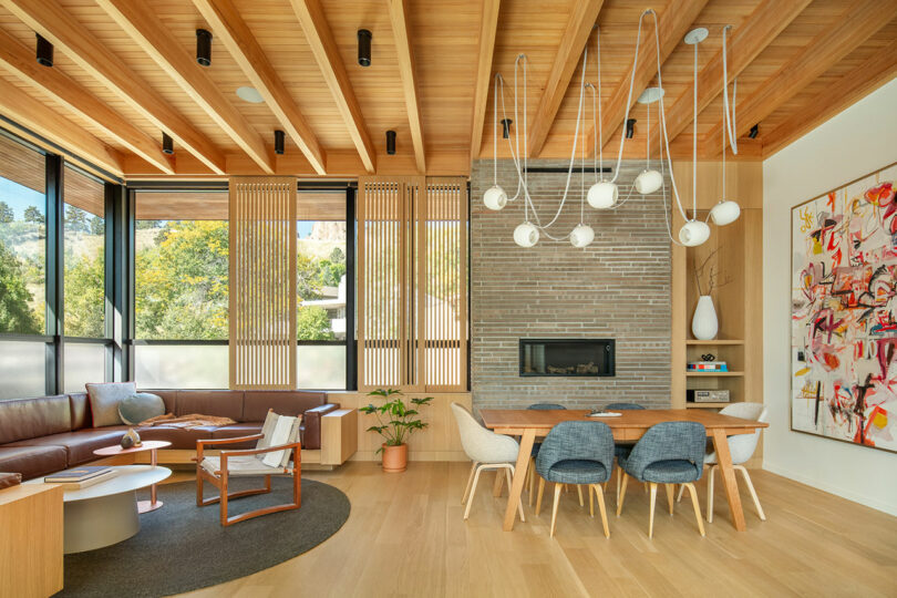 Modern living and dining area with wooden ceiling beams, large windows, sectional sofa, round coffee table, fireplace, dining table with blue chairs, abstract wall art, and a potted plant.