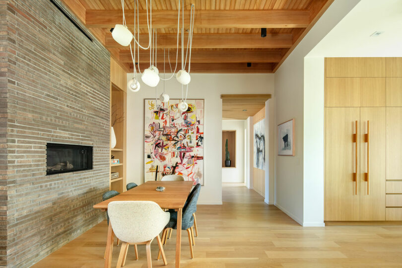 A modern dining room featuring a wooden dining table with mixed chairs, a brick wall with a fireplace, and a colorful painting. The room has wooden ceiling beams and light wooden flooring.