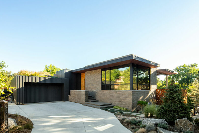 Modern house with large windows, brick and wood exterior, attached garage, and a concrete driveway surrounded by landscaping and greenery.