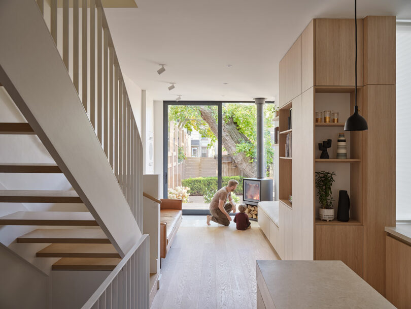 A person and a child sit on the floor in a bright, modern living room with light wood furniture and a large window showing a garden outside. An open staircase is on the left side of the room.