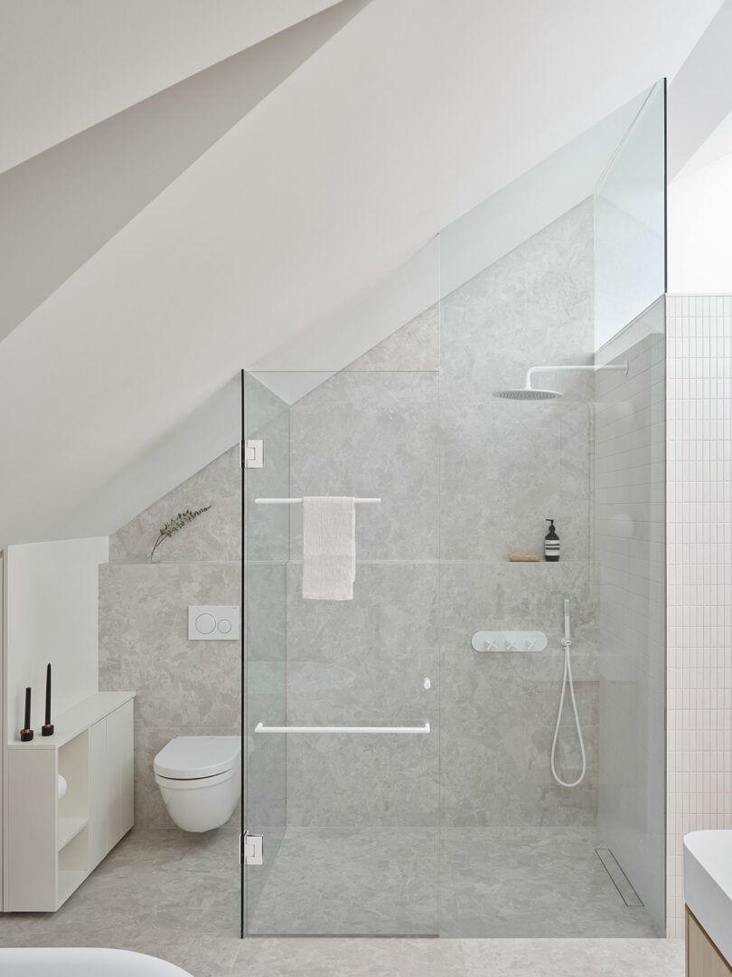 Modern bathroom with a glass-enclosed shower, wall-mounted toilet, and a sleek vanity. The shower features gray stone tiles, a built-in niche, and a rain showerhead. Two black candles stand on the vanity.