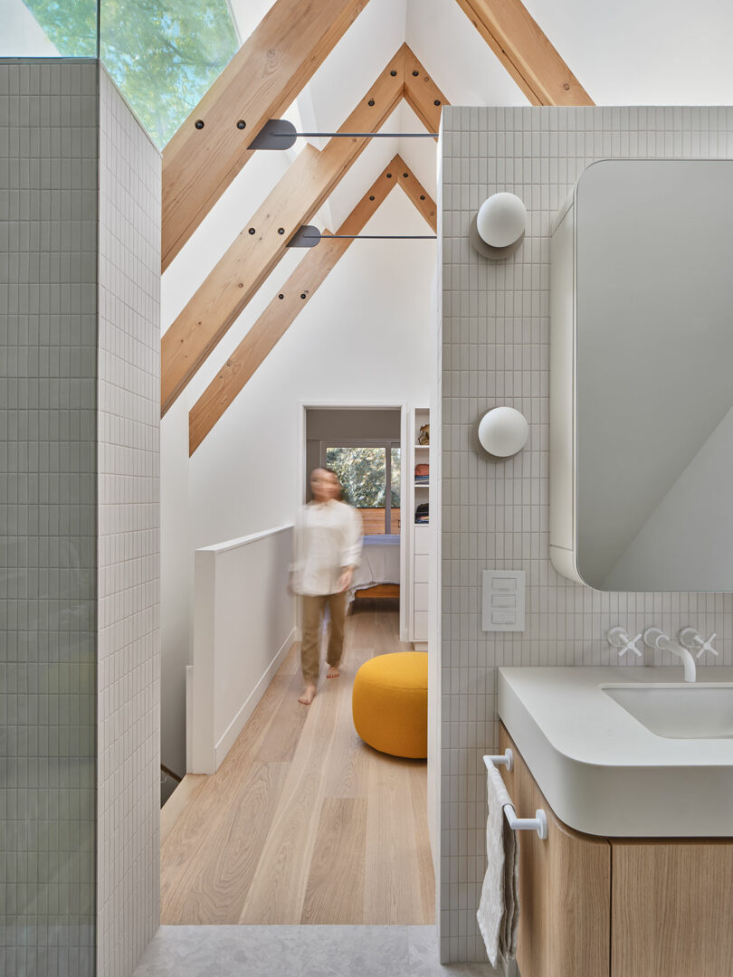 A person walks down a hallway with wooden beams overhead. The foreground features a modern bathroom with a large mirror, round lights, and a wall-mounted sink.