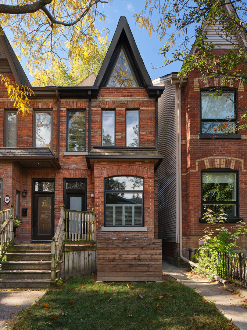 A two-story brick townhouse with a peaked roof and front steps, flanked by similar attached homes. Trees with autumn foliage are visible around the property.