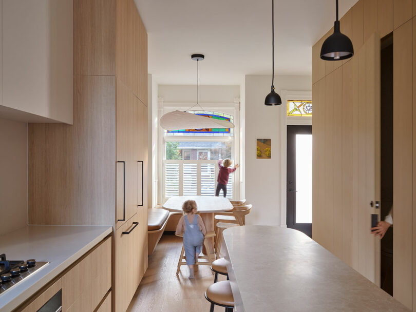 Two children in a contemporary kitchen, one standing on a bench near a window with stained glass and the other wearing overalls near a kitchen island. An adult's hand is partially visible near a door.