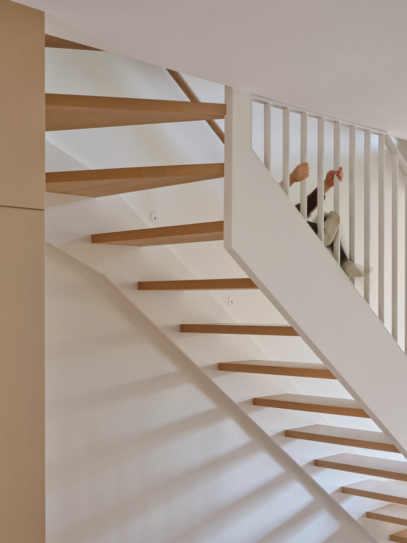 A modern staircase with wooden steps and white vertical rails, viewed from below. Two legs in white pants are visible through the railing.