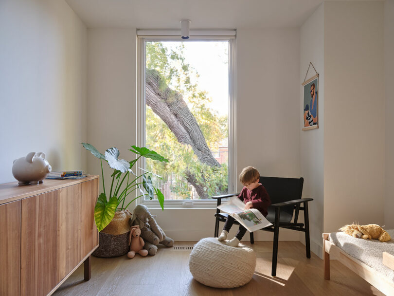 A child sits on a black chair reading a book in a bright room with a large window. A wooden cabinet, a pouf, a plant, and stuffed animals are in the room.