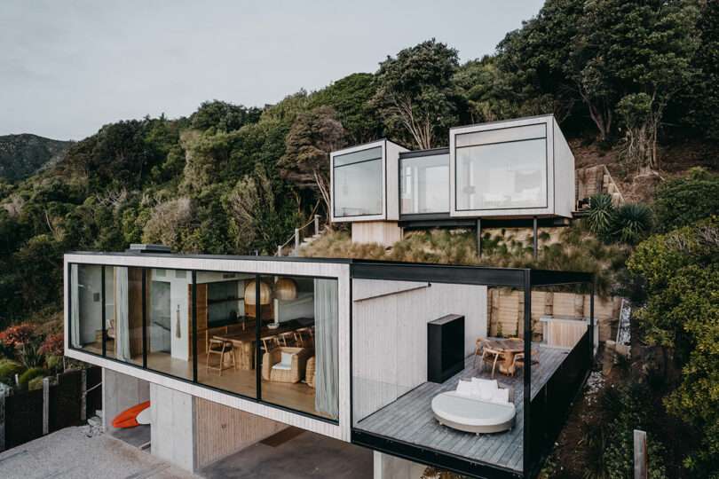A modern hillside house with large glass windows, featuring an open living space and separate elevated units surrounded by greenery. An outdoor seating area and greenery are visible.